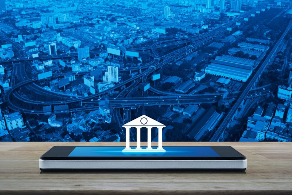 Smartphone com um ícone de um prédio bancário em seu display, sobre uma mesa de madeira. Ao fundo, uma paisagem urbana com prédios e uma rodovia, simbolizando a modernização e a conectividade dos bancos digitais.