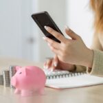 Mão usando o celular, com calculadora, cofrinho e pilhas de moedas ao lado. Simboliza o planejamento e a gestão financeira.