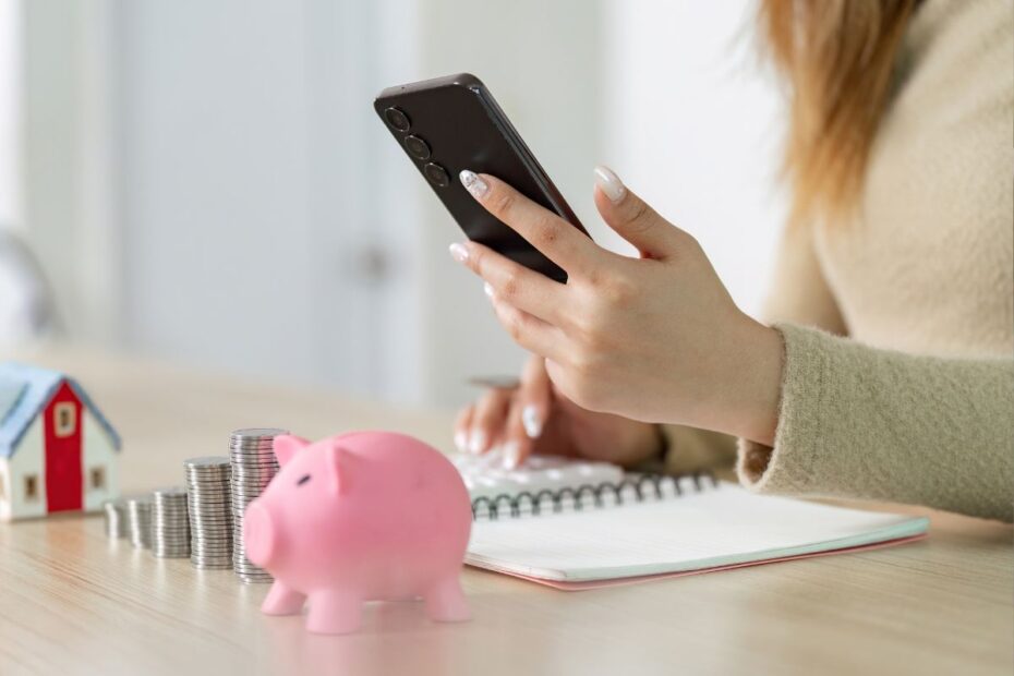 Mão usando o celular, com calculadora, cofrinho e pilhas de moedas ao lado. Simboliza o planejamento e a gestão financeira.