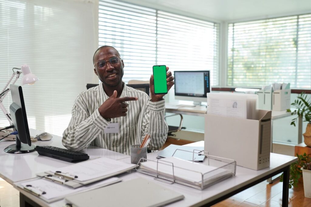 Homem negro sorrindo e apontando para um smartphone com a tela verde. Ele demonstra a alegria e o empoderamento de uma gestão financeira bem-sucedida.