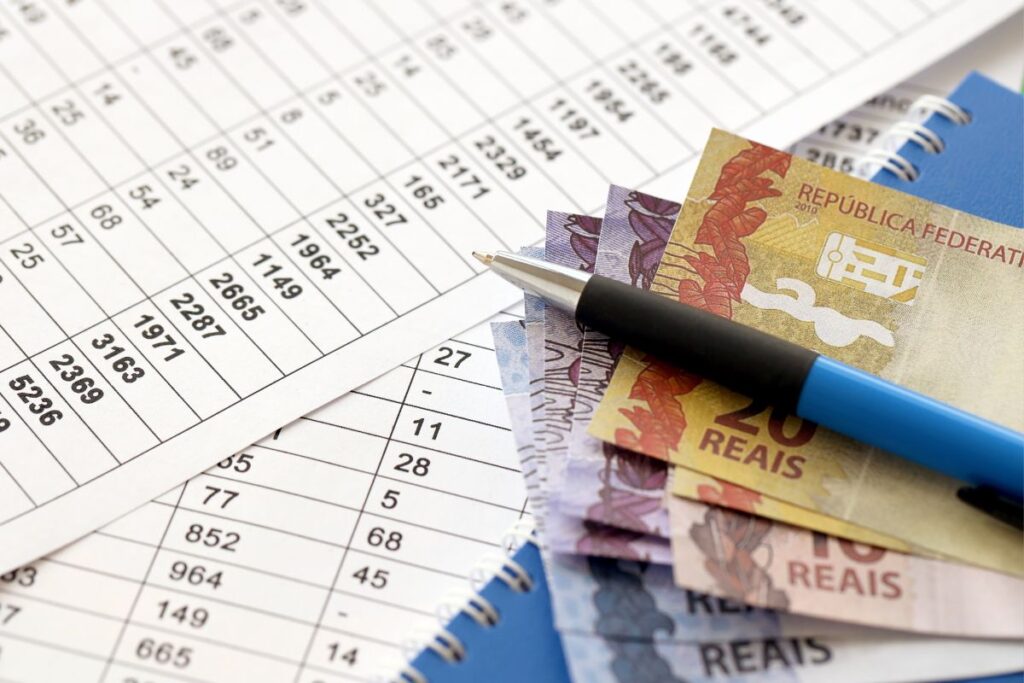 Planilha financeira com notas de real e caneta mostrando cálculos de benefícios