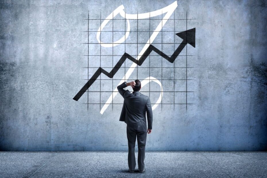 Homem de terno olhando para um gráfico de percentual (Juros) em ascensão na parede, representando o desafio das taxas elevadas.