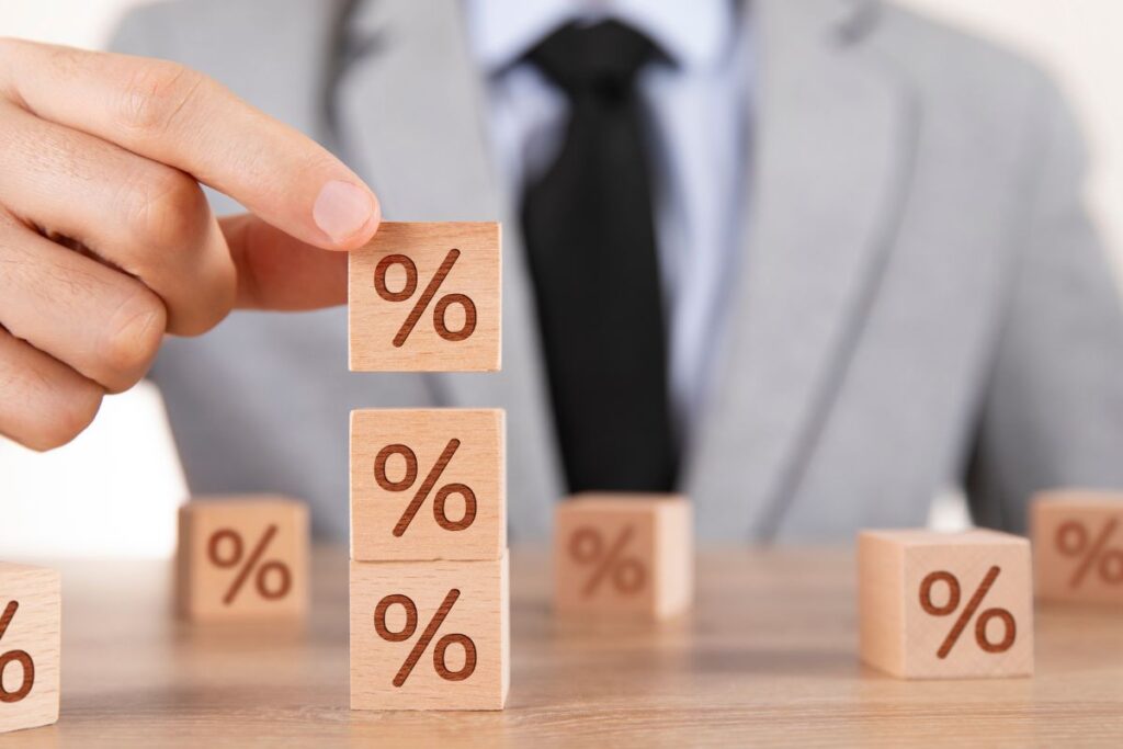 Mão de terno empilhando blocos de madeira com o símbolo de porcentagem (%), mostrando o controle sobre as taxas de juros pesados.