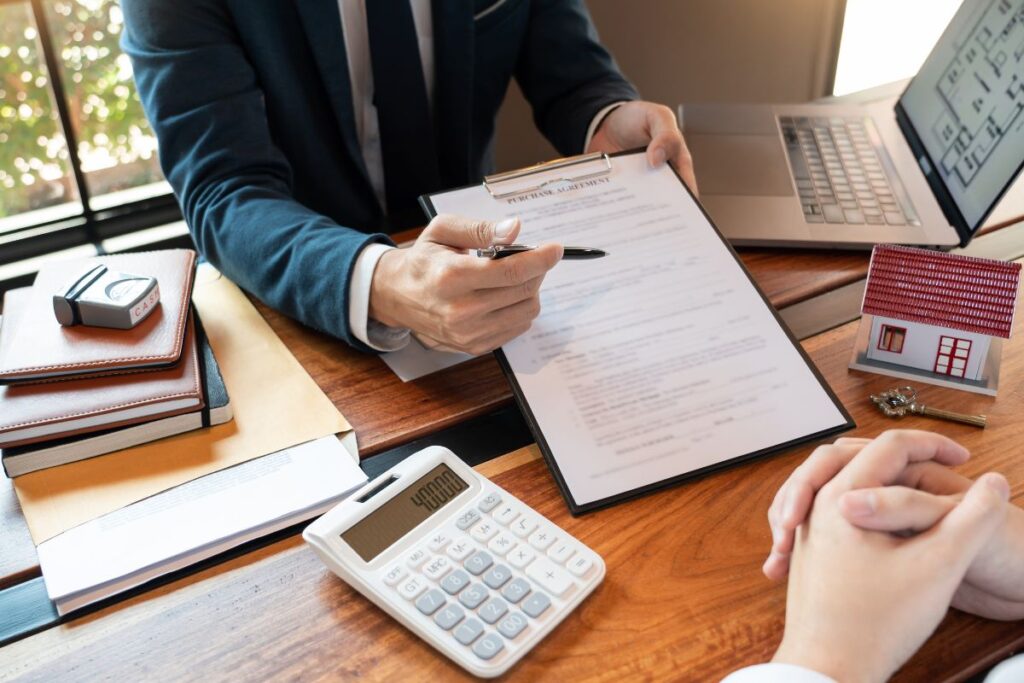 Consultor financeiro explica os termos de um contrato de empréstimo para um cliente, com uma calculadora e uma pequena casa de maquete na mesa. Representa a etapa de formalização de um crédito imobiliário.