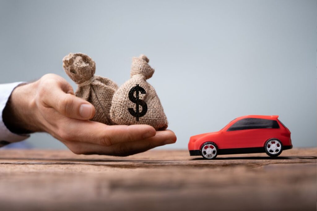 Uma mão segura dois sacos de dinheiro, com um símbolo de dólar, estendendo-os em direção a uma pequena maquete de um carro vermelho. Representa um financiamento de veículo ou empréstimo para compra de carro.