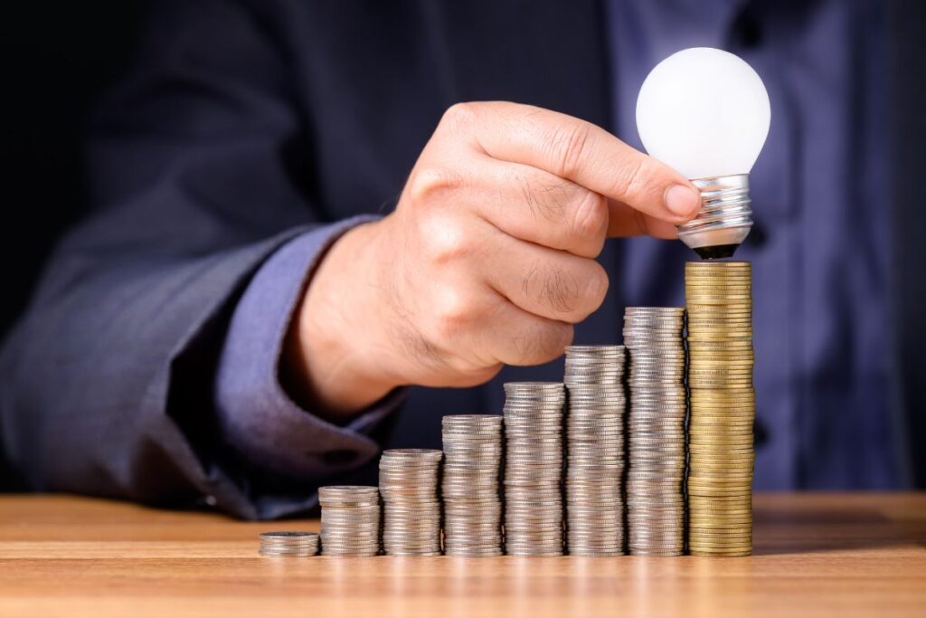 Uma mão segurando uma lâmpada acesa sobre uma pilha crescente de moedas, simbolizando a geração de ideias para o crescimento financeiro e a gestão de patrimônio.