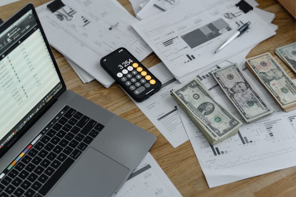 Mesa de trabalho com laptop, celular, relatórios financeiros e dinheiro para gestão de renda extra digital