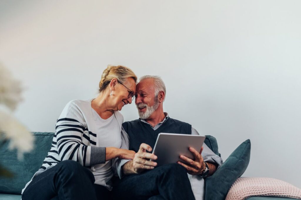Um casal de idosos sorrindo e olhando para um tablet. Eles estão sentados juntos em um sofá, mostrando a importância do planejamento financeiro conjunto para desfrutar da aposentadoria.