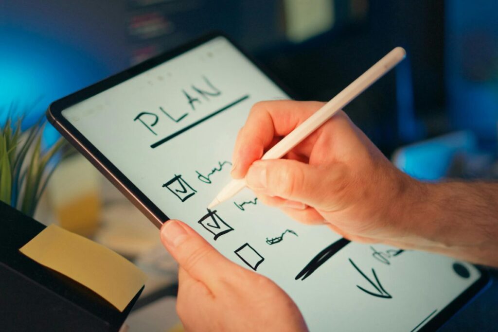 Uma mão usando uma caneta digital para checar itens em uma lista escrita à mão em um tablet, que tem a palavra "PLAN" no topo. A imagem simboliza o processo de planejamento de aposentadoria e metas financeiras.