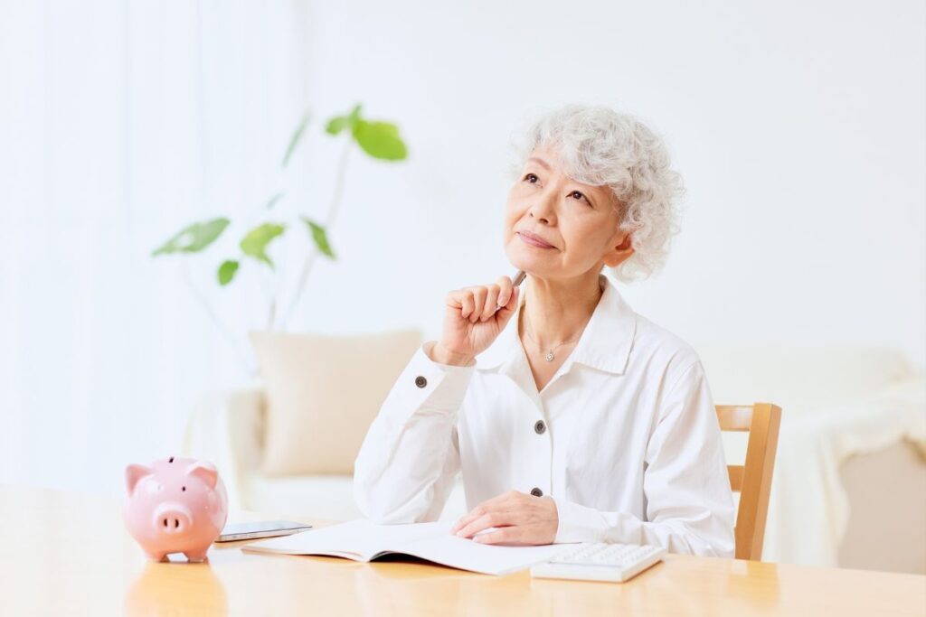 Uma mulher idosa, pensativa, sentada em uma mesa com um cofrinho de porquinho ao lado de um caderno aberto. A imagem representa o ato de ponderar sobre o planejamento de aposentadoria e o futuro financeiro.