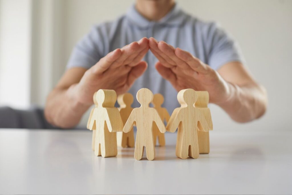 Representação de proteção através do seguro, com figuras humanas de madeira sendo protegidas por mãos em formato de cobertura.