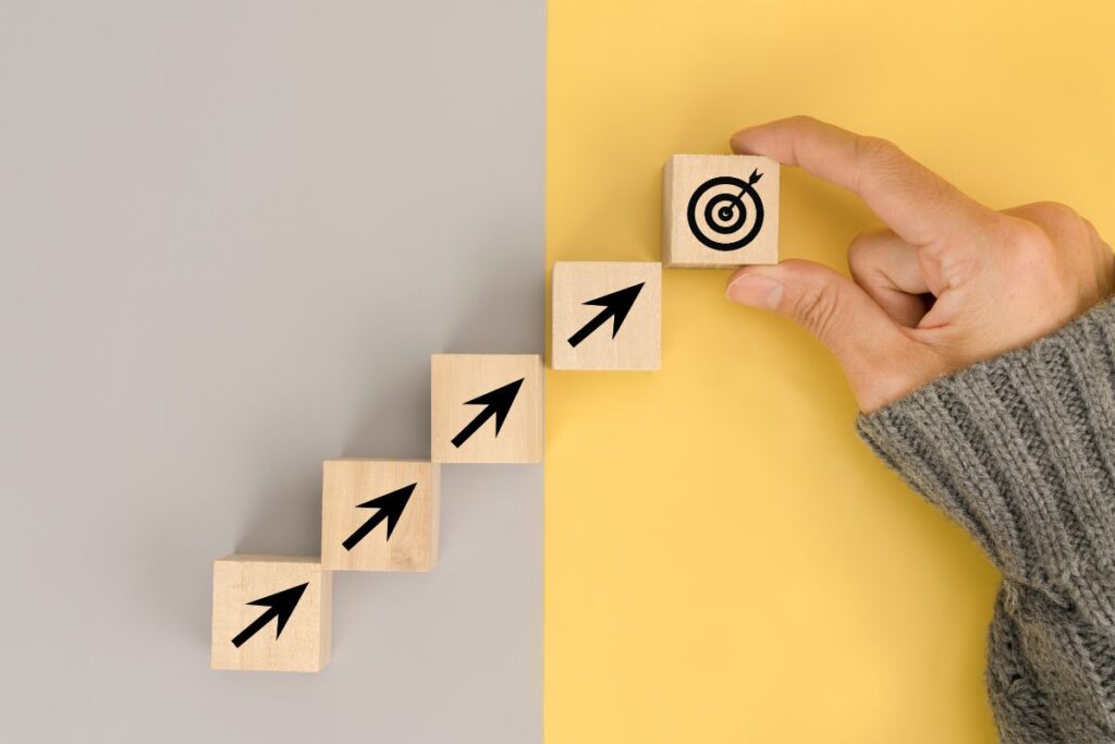 Uma mão posiciona um bloco de madeira com um alvo de meta no topo de uma escada ascendente de blocos com setas, simbolizando o progresso e o alcance de objetivos financeiros.