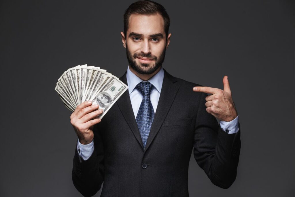 Homem de terno segurando leque de notas de dólar apontando para dinheiro simbolizando sucesso em construir.