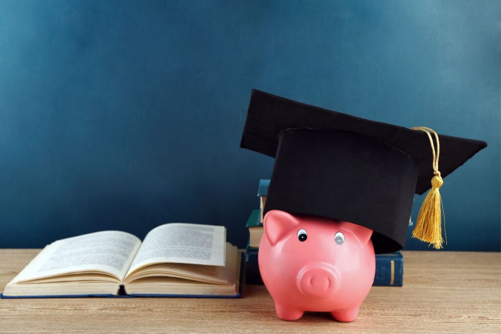 Cofre porquinho rosa com capelo de formatura ao lado de livros abertos simbolizando alfabetização sobre dinheiro