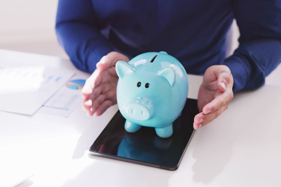 Um cofrinho azul de porquinho em cima de um tablet, sendo protegido pelas mãos de uma pessoa que o cerca com cuidado representando a previdência privada.