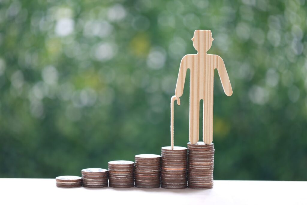 Um boneco de madeira, representando uma pessoa idosa com bengala, de pé sobre a pilha mais alta de moedas, que crescem em degraus sobre um fundo verde ao representar a previdência privada.