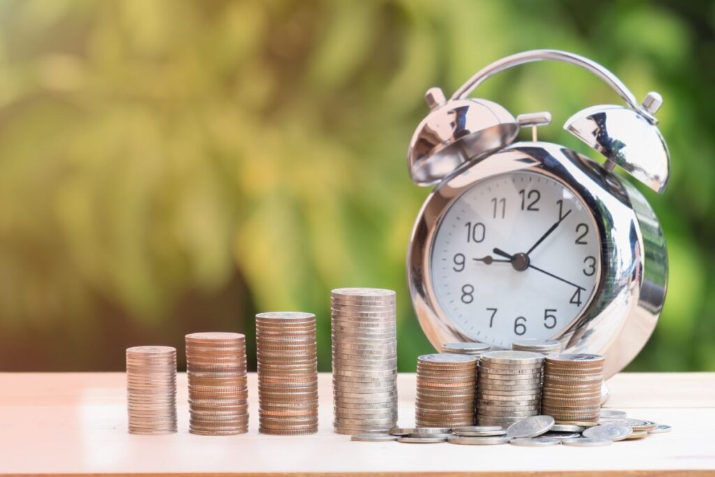 Pilhas crescentes de moedas de diferentes tamanhos, com um relógio despertador prateado ao fundo, em um cenário de luz natural e verde.