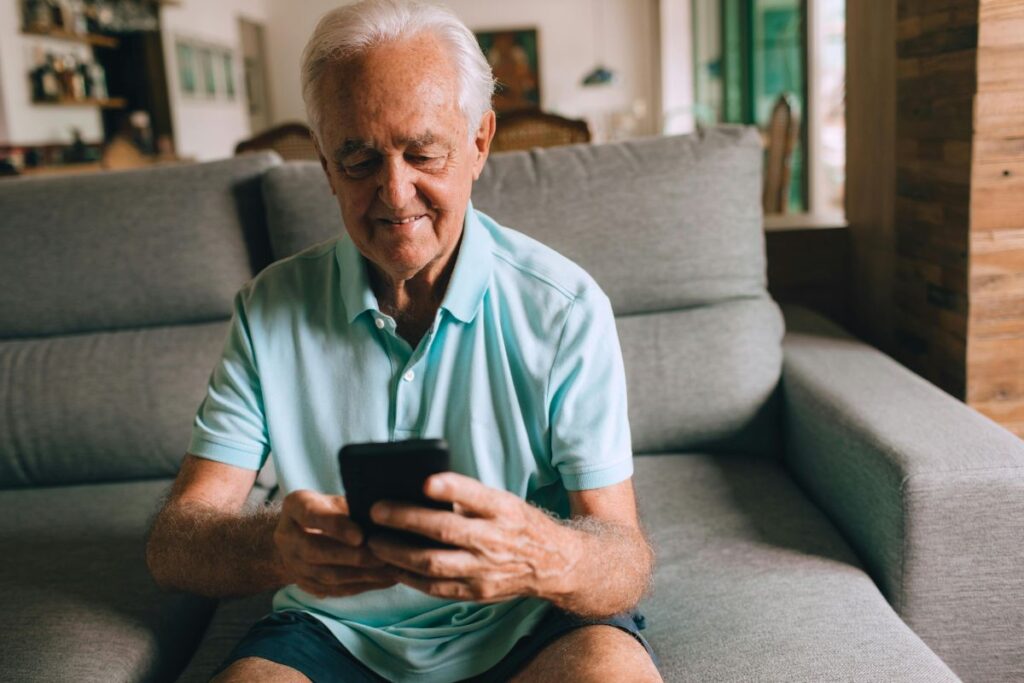 Um senhor idoso, com cabelos brancos, senta em um sofá e sorri gentilmente enquanto usa um smartphone, aparentemente consultando algo com satisfação.