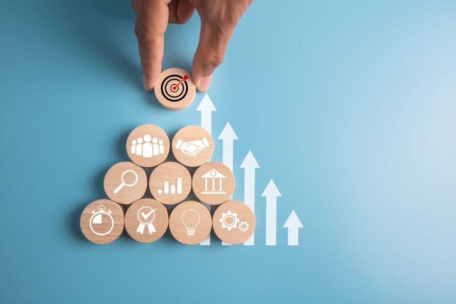 Mão colocando um bloco de madeira com um alvo no topo de uma pirâmide de blocos representando pilares (equipe, negociação, análise, etc.) e setas de crescimento, simbolizando metas e objetivos estratégicos.