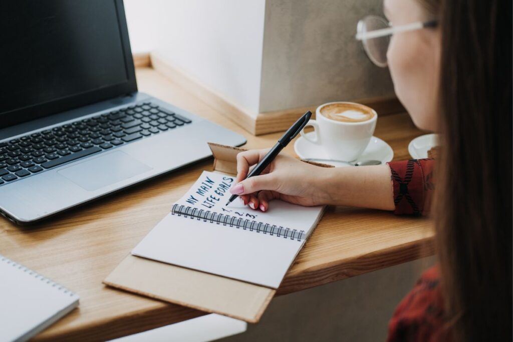 Pessoa de óculos escrevendo "MY MAIN LIFE GOALS" (Meus Principais Objetivos de Vida) em um caderno ao lado de um laptop e uma xícara de café, focando no planejamento de metas financeiras.