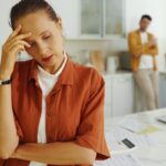 Mulher estressada na cozinha com a mão na testa e papéis sobre a mesa, refletindo a preocupação com novas dívidas ou contas.
