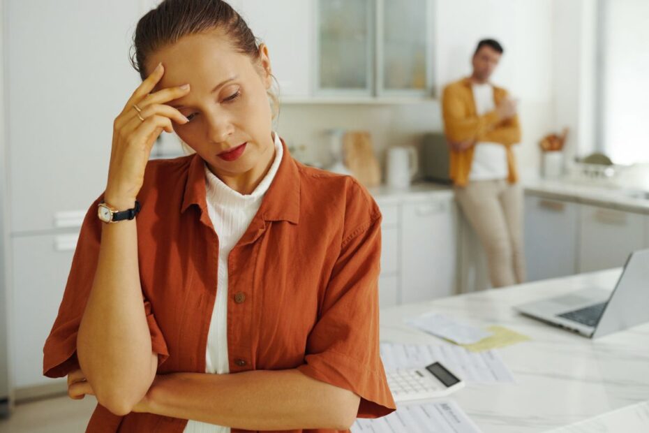 Mulher estressada na cozinha com a mão na testa e papéis sobre a mesa, refletindo a preocupação com novas dívidas ou contas.